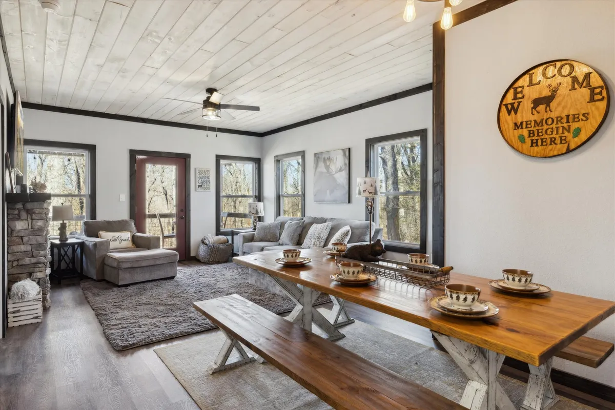 An inviting interior that seamlessly combines a dining table, comfortable seating area, and a fireplace. Large windows overlook the surrounding woods, creating a warm, communal atmosphere ideal for family meals and relaxing gatherings.