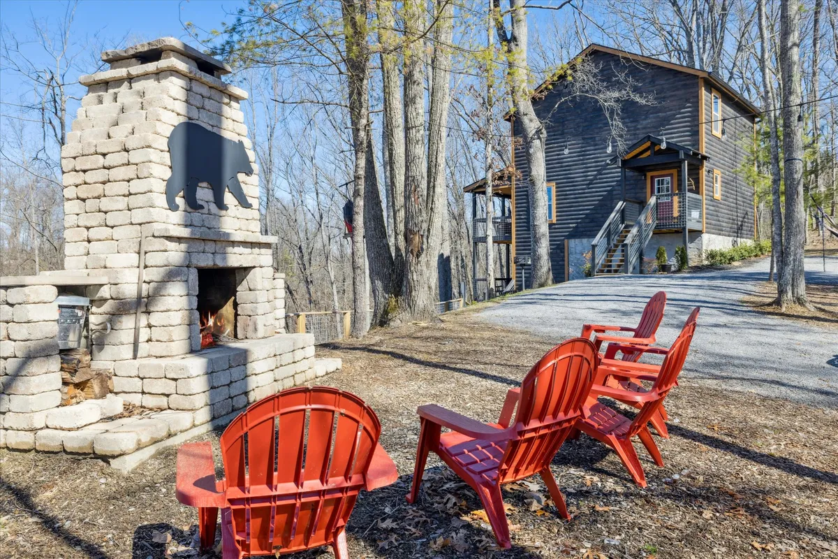 An inviting exterior space featuring a stone fireplace surrounded by red Adirondack chairs, set against wooded surroundings and a glimpse of a cabin-style house. The setting is ideal for evening gatherings, stargazing, and savoring the peaceful mountain atmosphere by the fire pit.