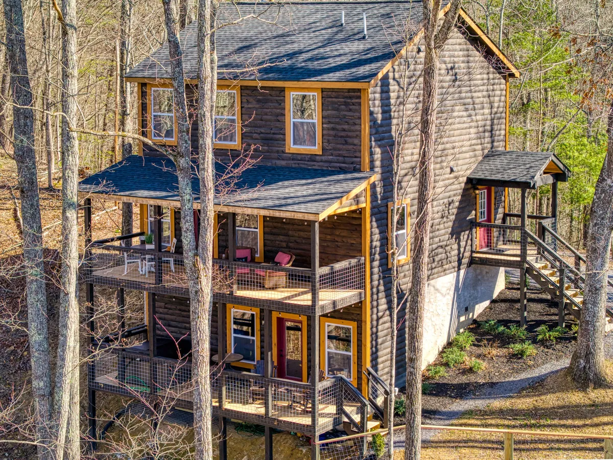 A three-story wooden cabin nestled among tall trees, featuring multiple balconies with chairs for relaxing. The setting is surrounded by nature, creating a sense of privacy and offering ample space for outdoor activities and gatherings.