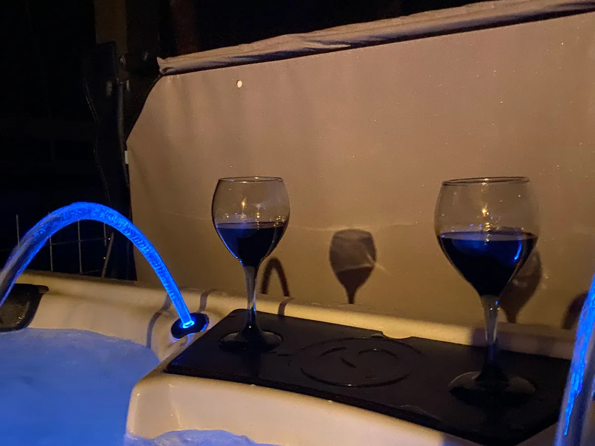 A close-up, atmospheric scene of two glasses of red wine perched on the edge of a bubbling hot tub. Subtle blue lighting and water features create an inviting ambiance, ideal for relaxing after a day of adventure.
