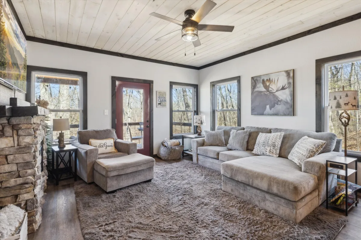 A spacious living room featuring a large sectional sofa arranged around a striking stone fireplace. The space is accented by a wooden ceiling, decorative artwork, and large windows that let in abundant natural light and frame scenic woodland views.
