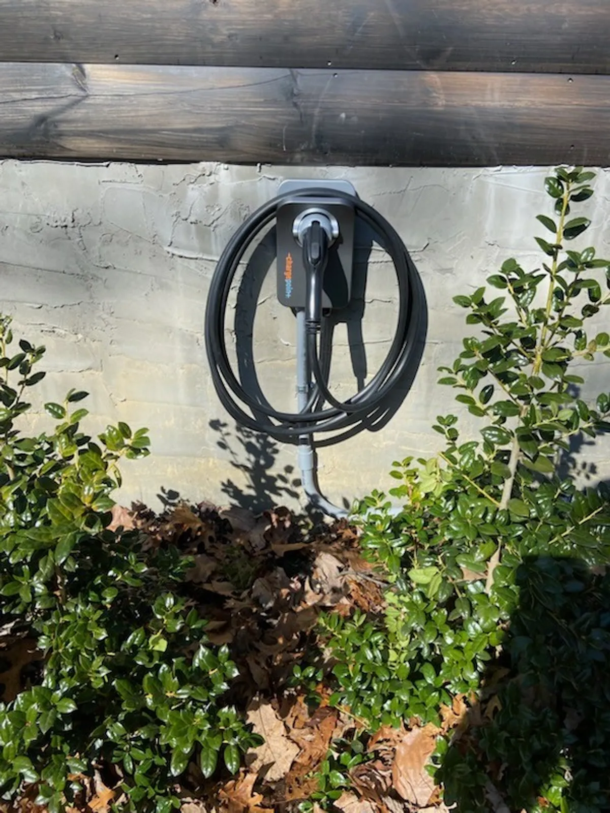 This exterior image centers on a sleek electric vehicle charging station nestled against a weathered wooden wall, surrounded by vibrant greenery. The scene seamlessly fuses modern technology with the tranquility of the mountain environment, highlighting a unique amenity directly relevant to guests looking to charge electric vehicles while exploring the area.