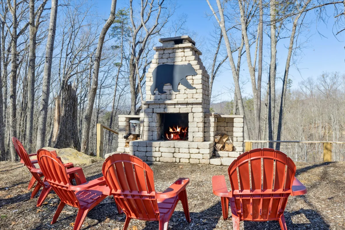 A stone fireplace with a bear silhouette is surrounded by red Adirondack chairs and set amidst tall trees. The warm glow and inviting seating evoke moments of gathering around the fire for stories and s’mores.