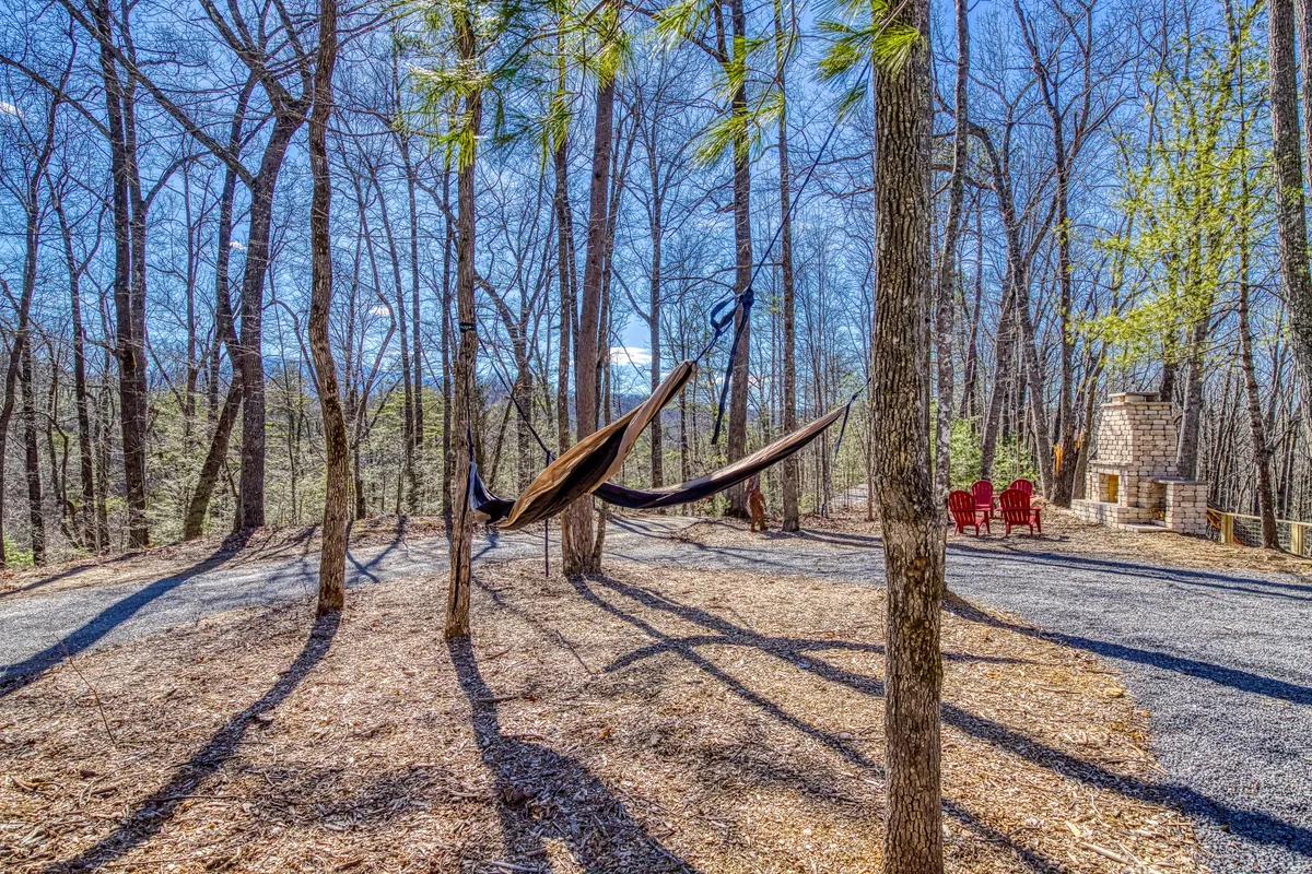 A hammock gently sways between trees near a rustic fireplace and red chairs, creating a tranquil nook for relaxation. The image conveys restful moments and the integration of leisure spaces in the natural setting.
