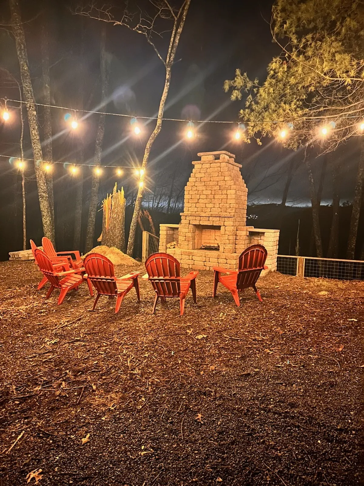 A cozy outdoor fire pit area with red Adirondack chairs and a brick fireplace, illuminated by string lights. The setup is perfect for storytelling, sharing laughter, and making memories under the stars.