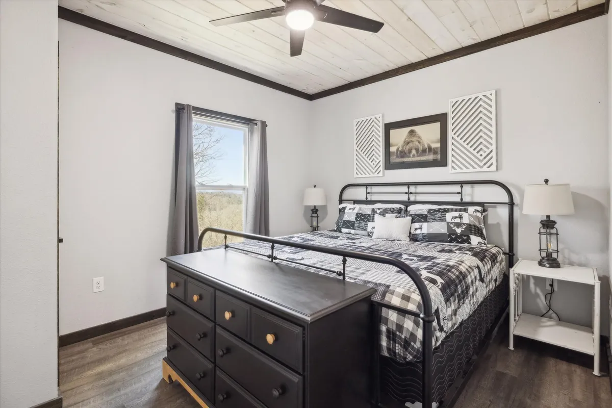 A serene bedroom featuring a bed with black and white bedding, a decorative wall art piece above the headboard, and a window offering tranquil views of the surrounding forested property. Soft natural light filters through the window, illuminating the calm and restful atmosphere perfect for unwinding.