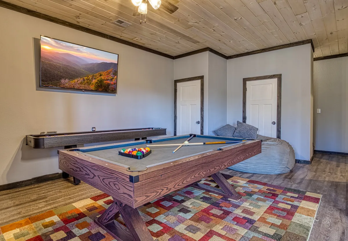 A lively game room setting with a pool table, a bean bag chair, a colorful rug, and a television displaying a scenic landscape. The space exudes a casual, fun atmosphere suitable for both games and relaxation.