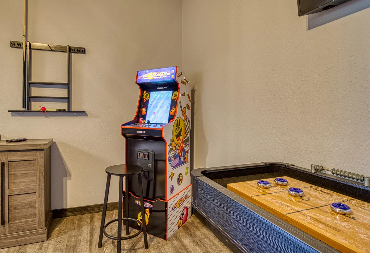 This image focuses on classic arcade entertainment, featuring an arcade game machine, a shuffleboard table, a stool, and wall shelving. The ambiance is playful and nostalgic, inviting guests to enjoy friendly competitions and memorable gatherings.