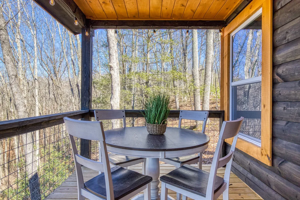 A welcoming outdoor dining area with a wooden table and white chairs set under the trees, accented by string lights and large windows in the background. The area invites guests to enjoy meals, morning coffee, or evening gatherings in nature.